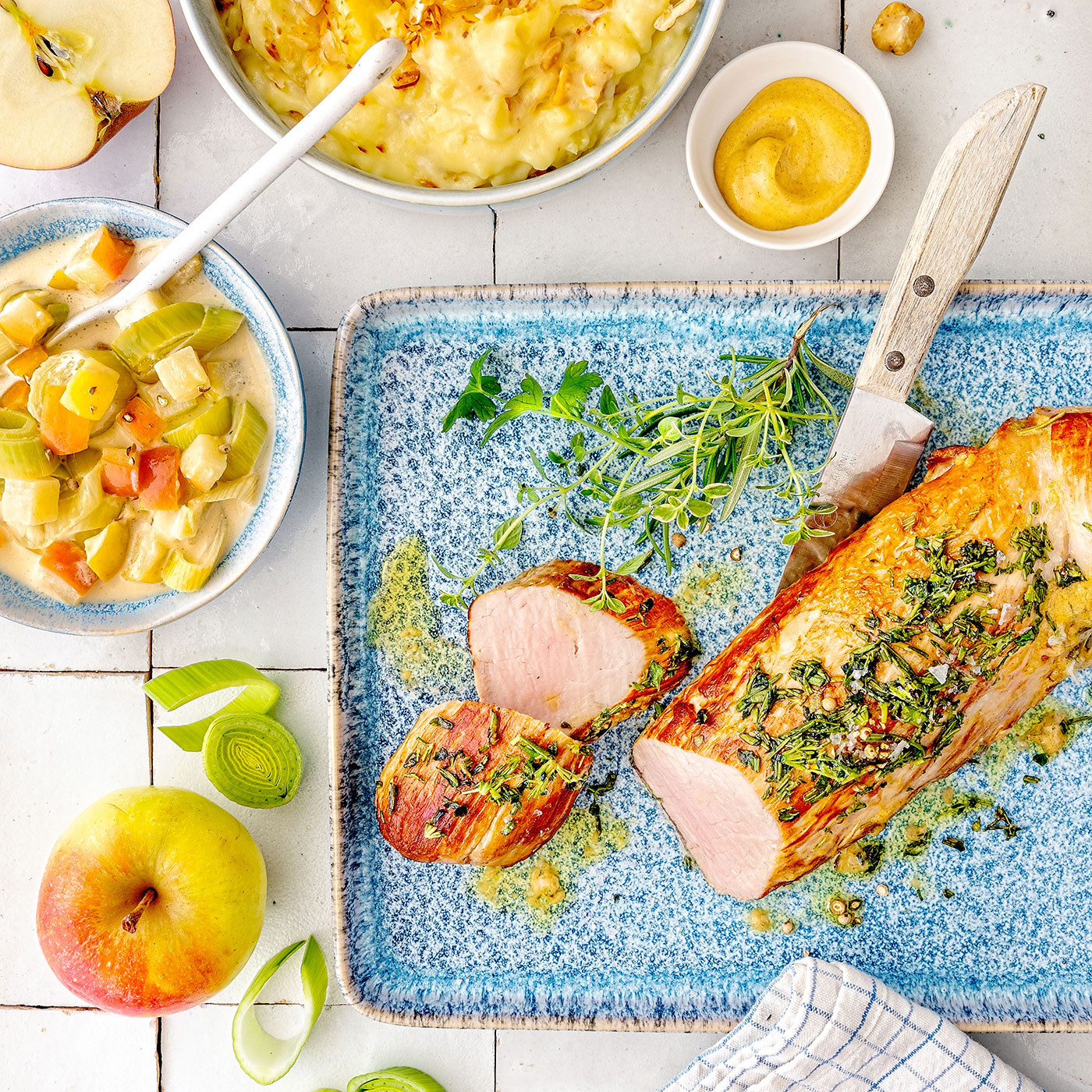 Schweinefilet im Winterkräutermantel mit Apfelgemüse, Kartoffelgratin und frischen Kräutern auf blauer Keramikplatte.