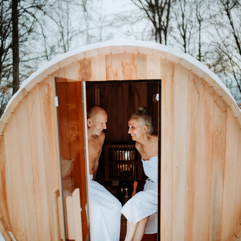 Zwei Menschen sitzen lachend in einer Außensauna im Schnee und genießen die winterliche Ruhe.