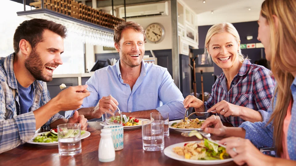 Zwei lachende Männer und zwei lachende Frauen essen gemeinsam Salat an einem brauen Holztisch. 
