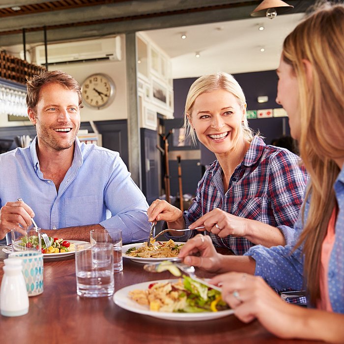 Zwei lachende Männer und zwei lachende Frauen essen gemeinsam Salat an einem brauen Holztisch. 