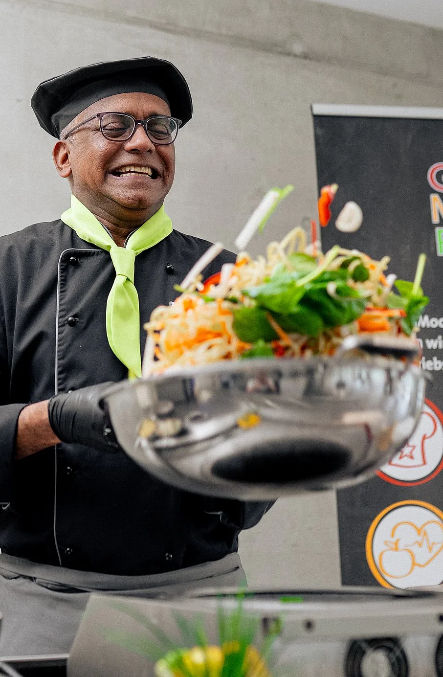 Koch, der einen Wok schwenkt. Rechts im Hintergrund steht ein Aufsteller auf dem "GOOD MOOD FOOD" steht