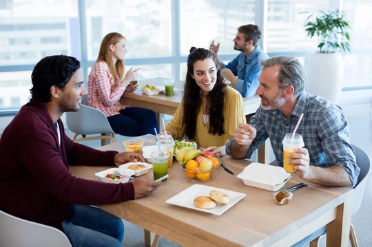 Im Vordergrund sitzen zwei Männer und eine Frau an einem Holztisch in einer Kantine. Auf dem Tisch liegen verschiedene Speisen und Smoothies. Im Hintergrund unterhalten sich Mann und Frau an einem weiteren Tisch.