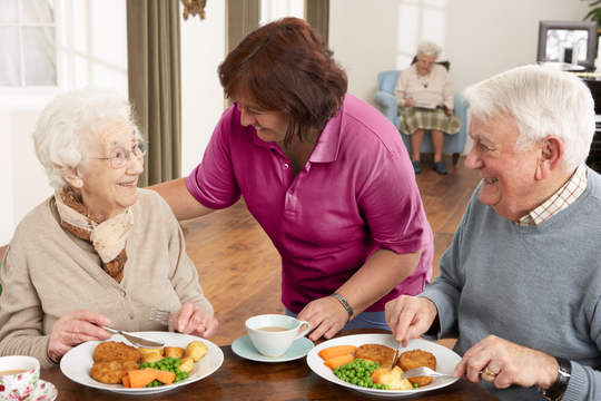 Zwei Senioren sitzen vor gesundem und bedarfsgerechten Essen und werden von einer Pflegekraft zum Mittag bedient