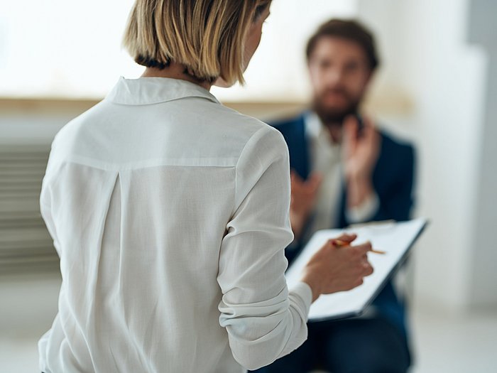 Mittig eine Frau in weißer Bluse, in ihrer Hand ein Stift und Checkliste. Im Hintergrund ein Mann, welcher versucht etwas zu erklären. 