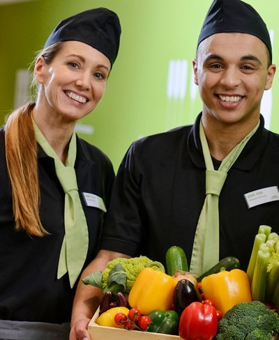 Zwei Mitarbeiter von apetito catering haben Spaß bei Ihrer Arbeit und präsentieren eine bunte Box mit vielseitigem frischen Gemüse