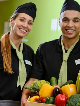 Zwei Mitarbeiter von apetito catering haben Spaß bei Ihrer Arbeit und präsentieren eine bunte Box mit vielseitigem frischen Gemüse