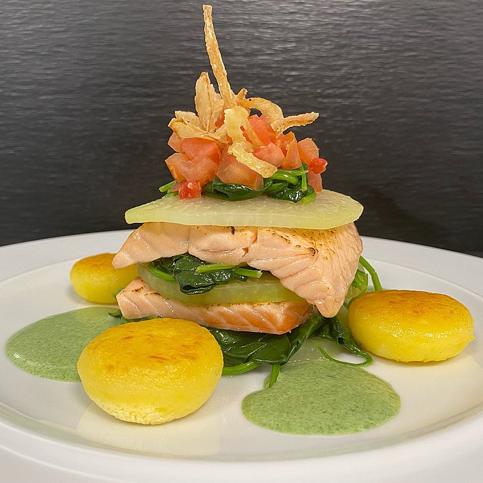 angerichtetes Gericht mit Lachsfilet, Blattspinat und dünnen Kartoffelscheiben, getoppt mit Tomatenwürfeln und Zwiebeln auf einem weißen Teller mit grüner Sauce