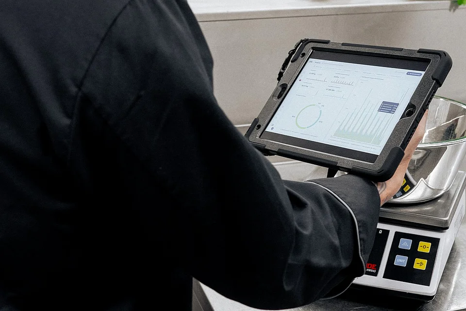 Ein Koch in schwarzer Kochjacke wiegt eine Schüssel und bedient dabei ein schwarzes Ipad.