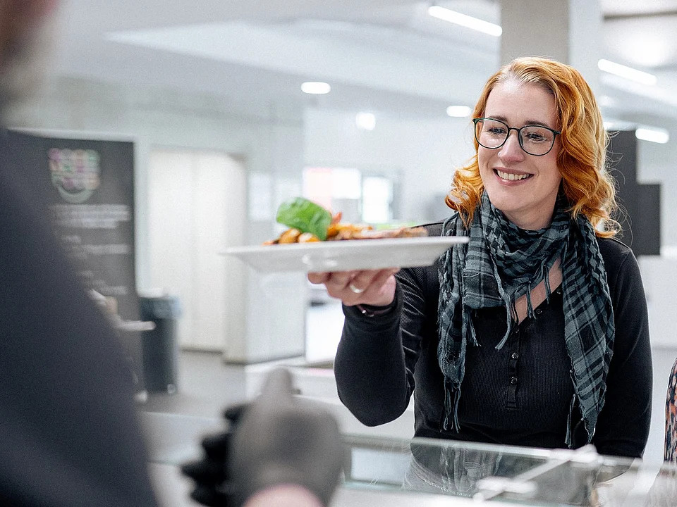 Drei Gäste stehen vor der Essenausgabe. Einer Frau wird ihr Essen überreicht.