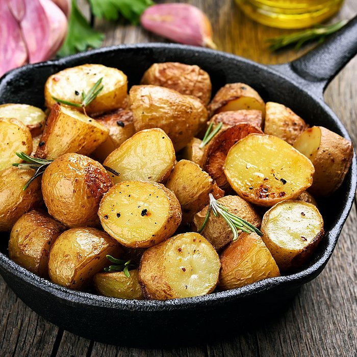 Rosmarinbratkartoffeln in einer Pfanne auf einem Holztisch. Im Hintergrund Knoblauchzehen und Öl
